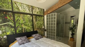 A bedroom with a large bed, a view of greenery through windows, a hanging light fixture, and a glass shower enclosure. - Bed and breakfast in the south of France (Entre-Vignes)
