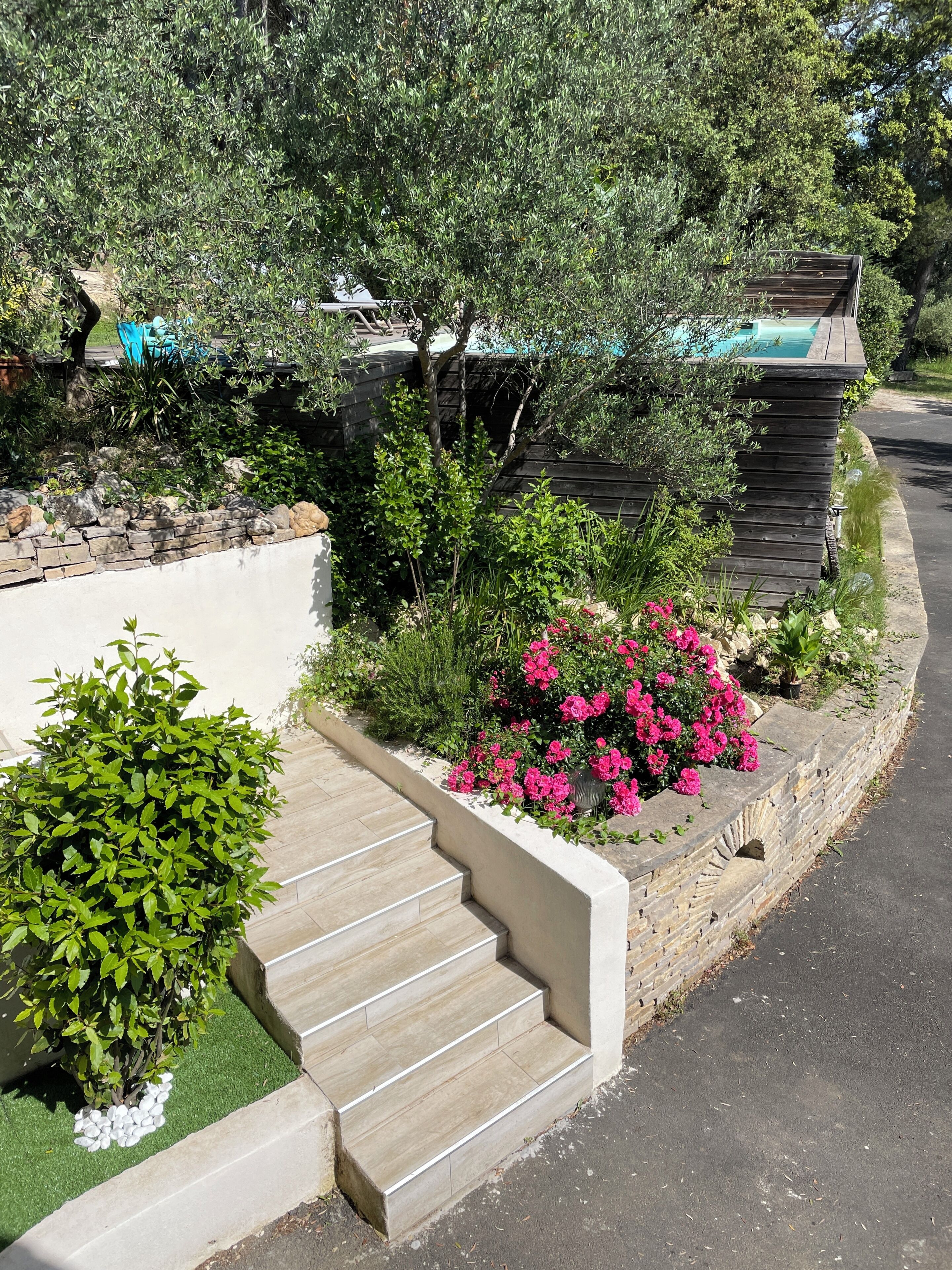 Un área ajardinada con un muro de piedra, escalones que llevan a una terraza de madera y una piscina.