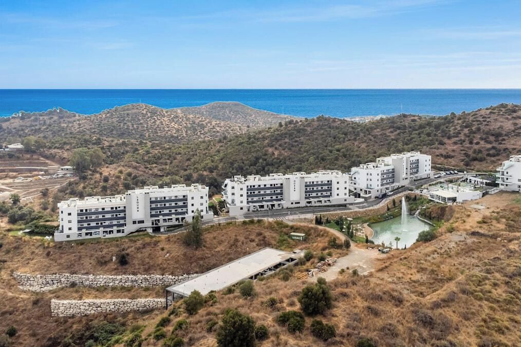 Un complexe hôtelier moderne composé de plusieurs bâtiments, doté d’un espace paysager agrémenté d’une fontaine, et offrant une vue sur la mer et les collines en arrière-plan.