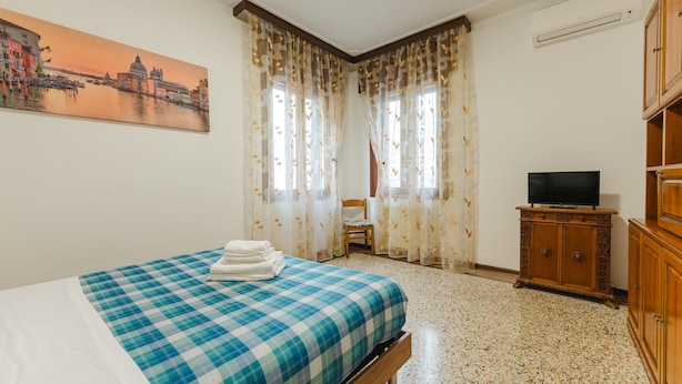 A hotel room with a bed, a television on a wooden cabinet, a chair, and a painting of a cityscape. - Casa Tatiana (Mestre)