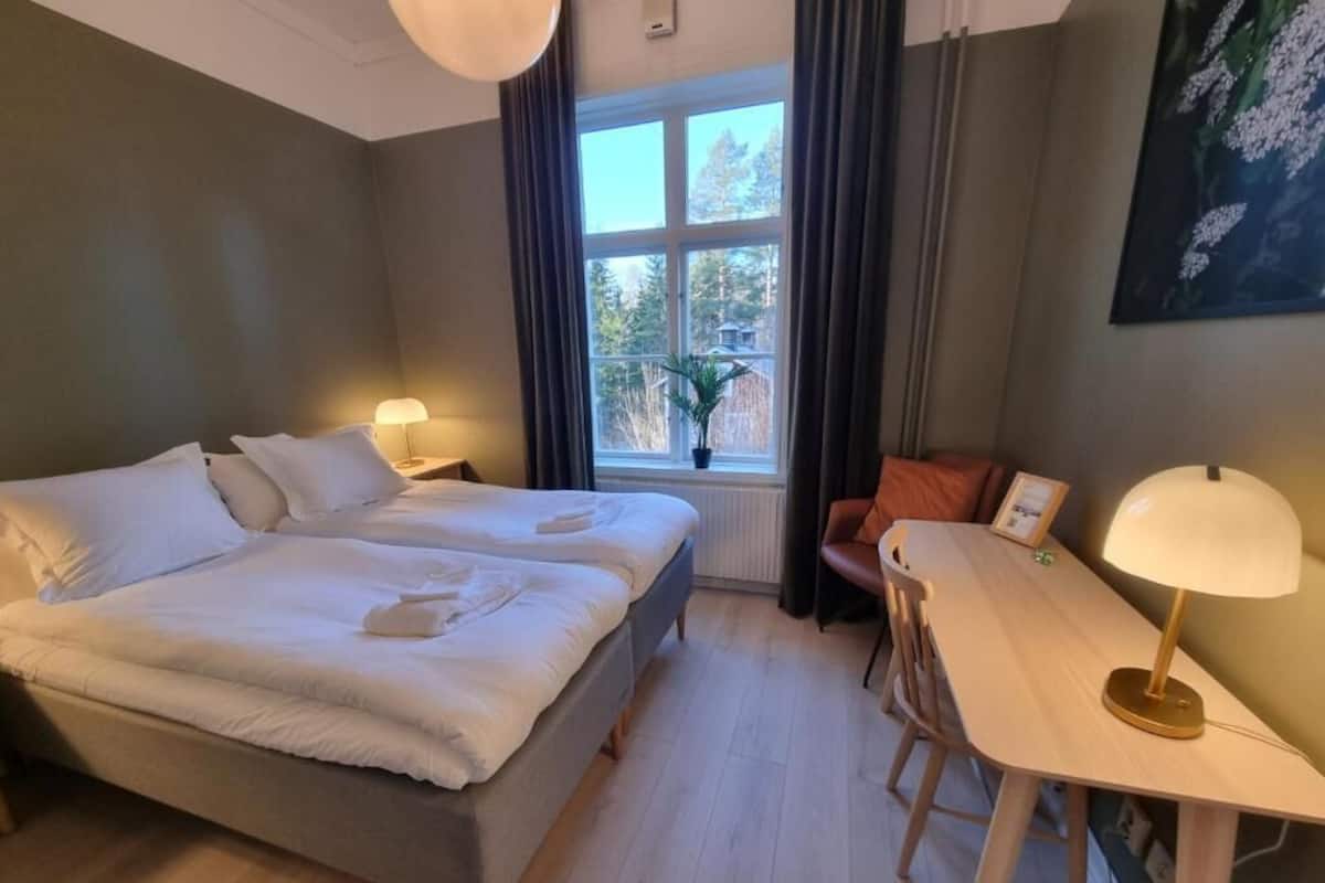 Une chambre à coucher avec un lit double, une petite table, une chaise, une fenêtre avec des rideaux et une plante en pot.