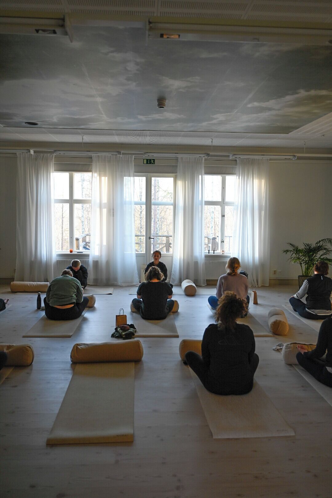 A room with large windows, a ceiling fan, and people sitting on mats.