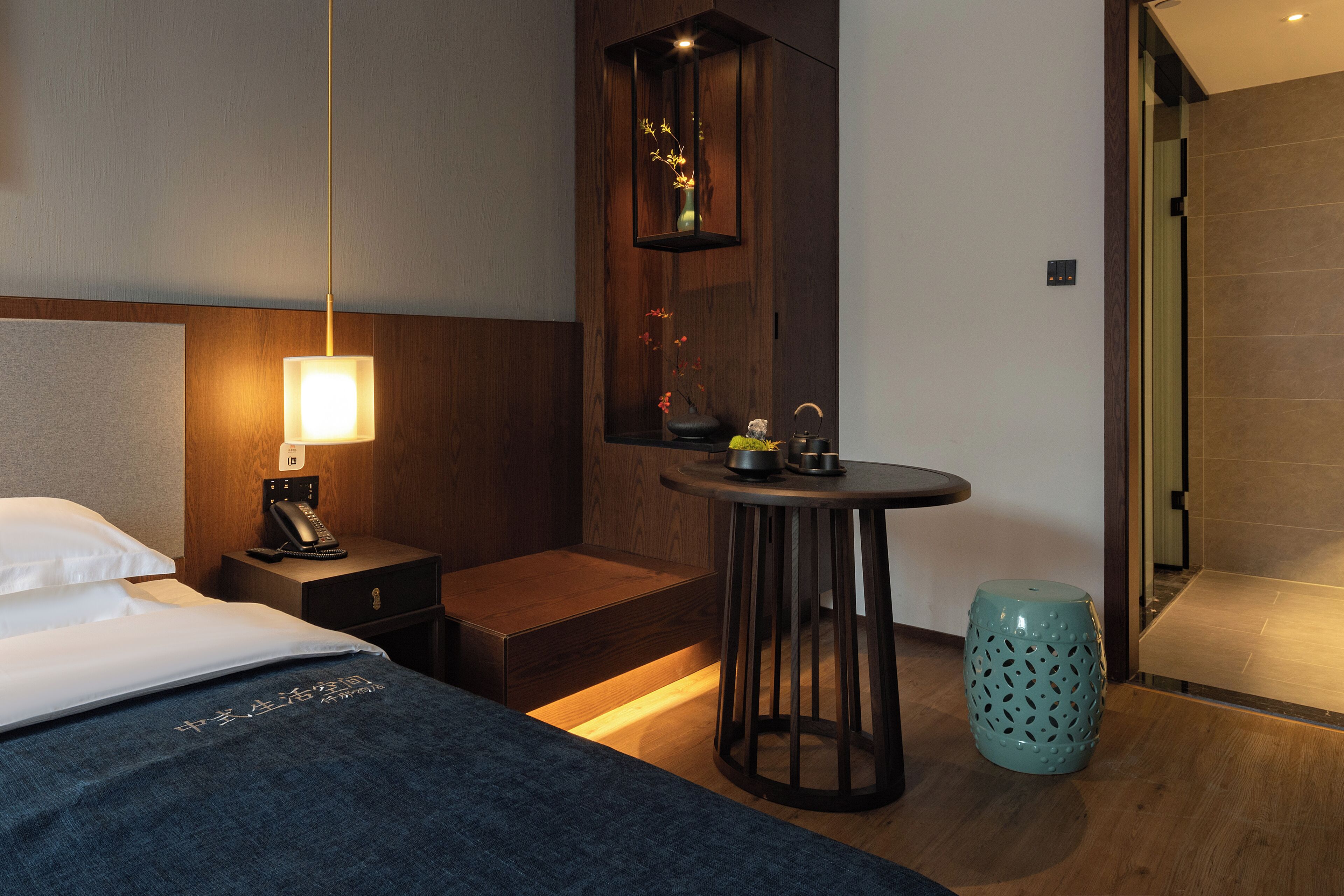 A hotel room with a bed, bedside table, lamp, small table with a vase, and a trash bin.
