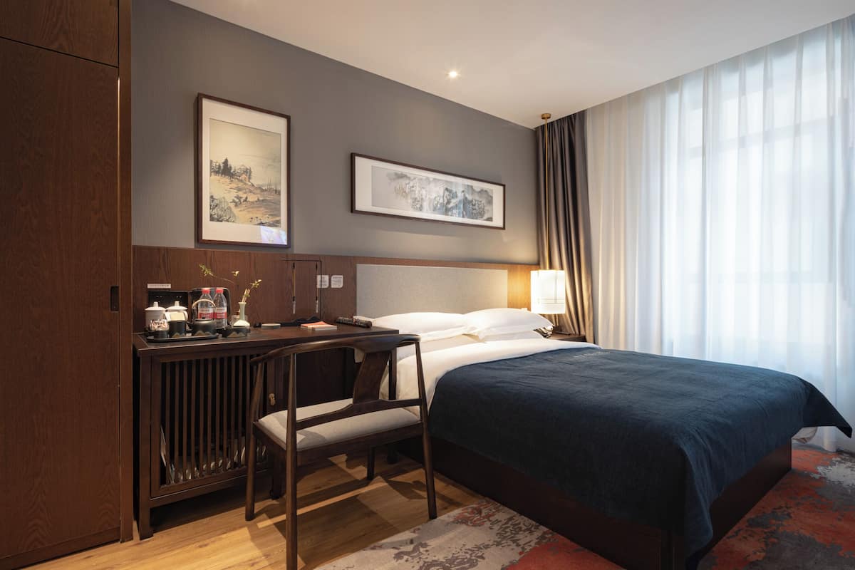 A hotel room with a bed, a desk with a chair, a lamp, and two framed pictures on the wall.