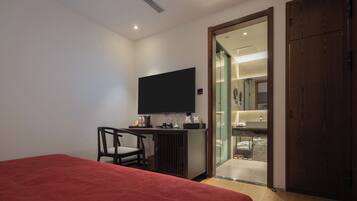 A bedroom with a bed, a desk, a chair, a TV, and a bathroom visible through a glass door.