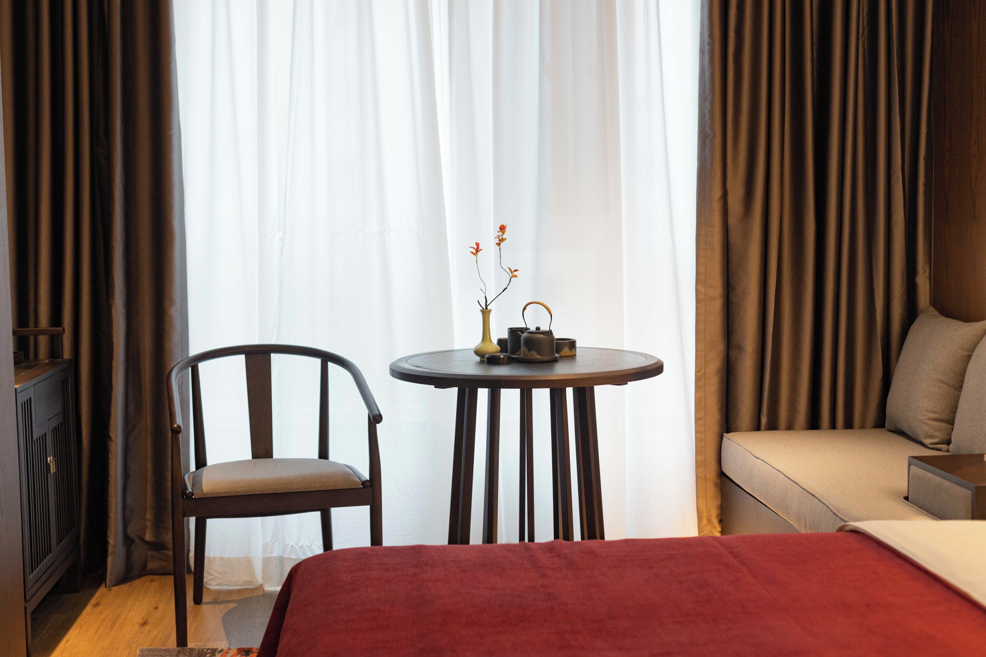 Una habitación de hotel con una cama, una silla, una mesita con un jarrón y una tetera, y un sofá.