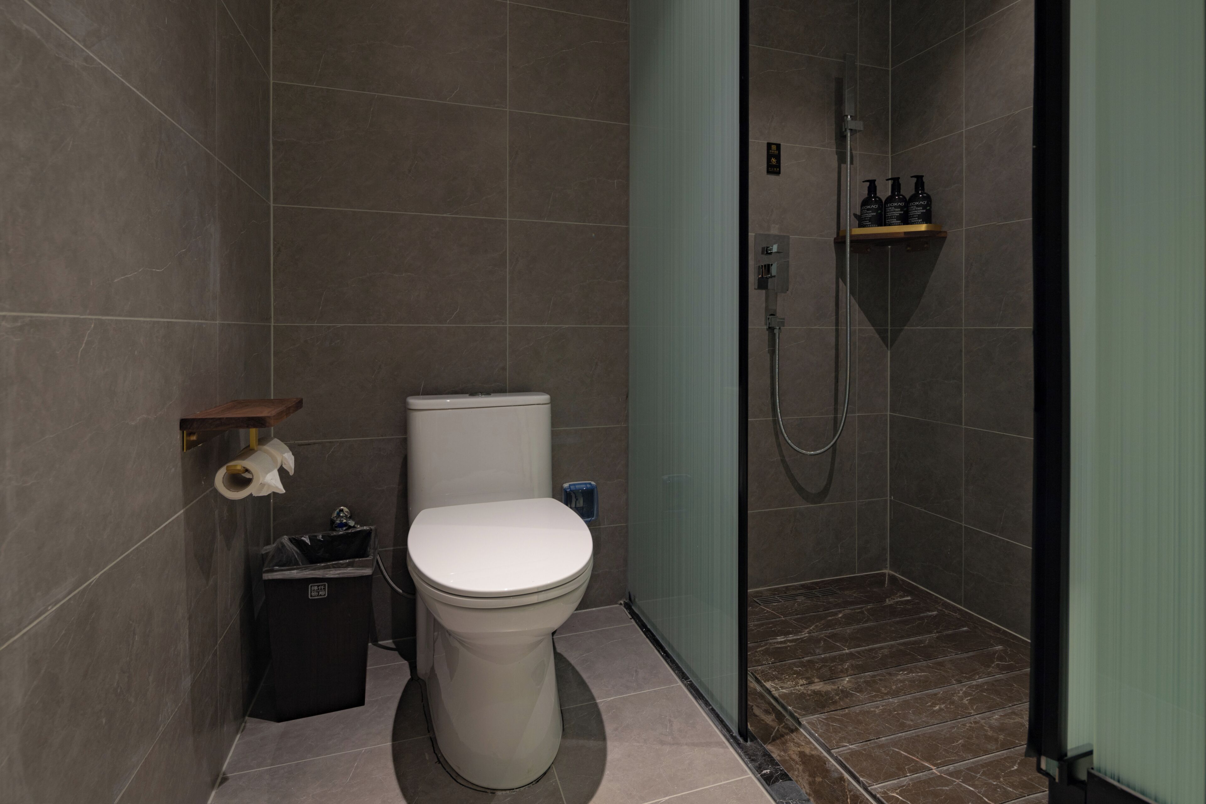 A modern bathroom with a toilet, a trash bin, a shower area, and a shelf with toiletries.