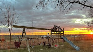 Un terrain de jeux avec des balançoires et un toboggan, entouré d’un champ d’herbe et d’arbres.