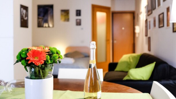 Ein Zimmer mit einem Bett, einem Tisch mit einer Flasche und Blumen, einer Couch mit Kissen und gerahmten Bildern an den Wänden.