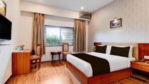 Een hotelkamer met een bed, bureau, stoel, klein tafeltje, raam met gordijnen, airconditioning en een lijst met foto aan de muur.