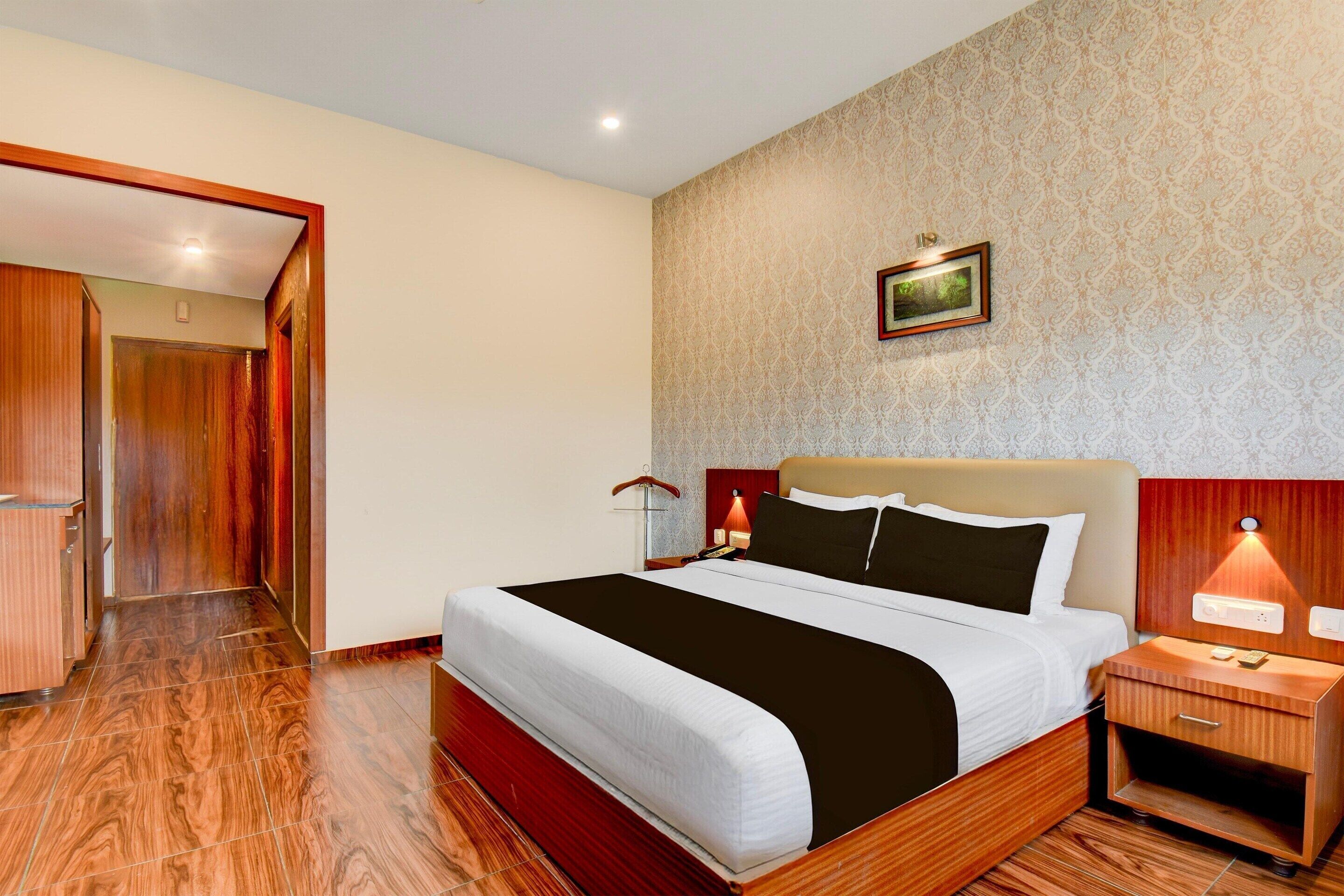 A hotel room with a wooden floor, a bed with a white and black bedspread, a nightstand with a lamp, and a framed picture on the wall.