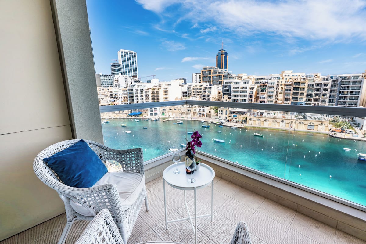 A balcony with a view of a marina and cityscape, a wicker chair, a small table with a bottle and glass, and a decorative flower.