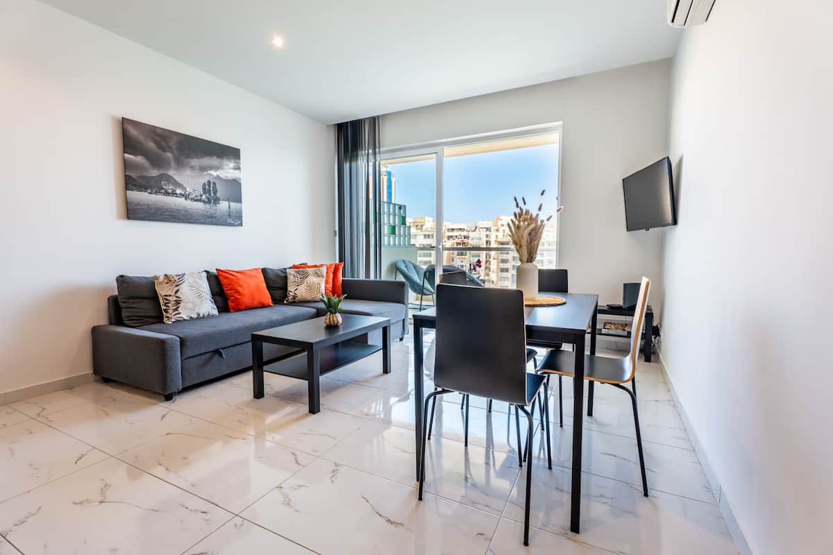 A modern living room with a sofa, coffee table, dining table, and chairs. There is a large window with a view of buildings and a flat-screen TV mounted on the wall.