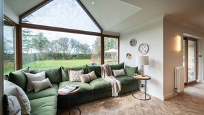 A green sofa with a side table, a lamp, and a view of a garden through large windows. - Luxury Golf House near Royal Birkdale (The Open) | Hot Tub | Sleeps 12+ (Southport)