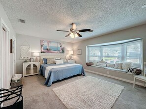 Ein Schlafzimmer mit einem Bett, Deckenventilator, Fenster mit Jalousien, Nachttisch und Wandregal.