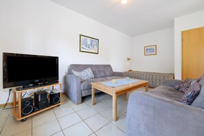Ein Wohnzimmer mit einem Flachbildfernseher, zwei Lautsprechern, einem grauen Sofa, einem Holztisch und zwei gerahmten Bildern an der Wand.