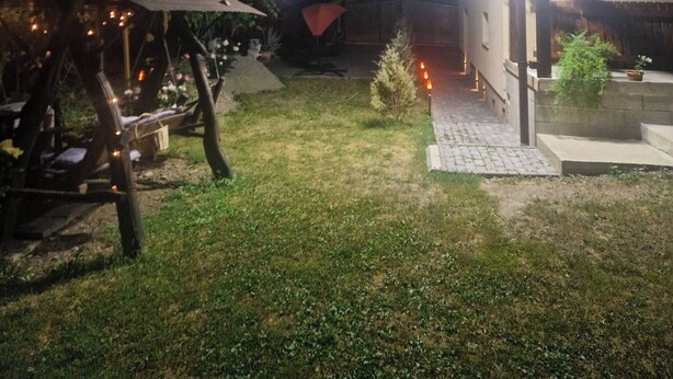 Ein Außenbereich bei Nacht mit einer Holzkonstruktion, einem Weg und einem Gebäude im Hintergrund.
