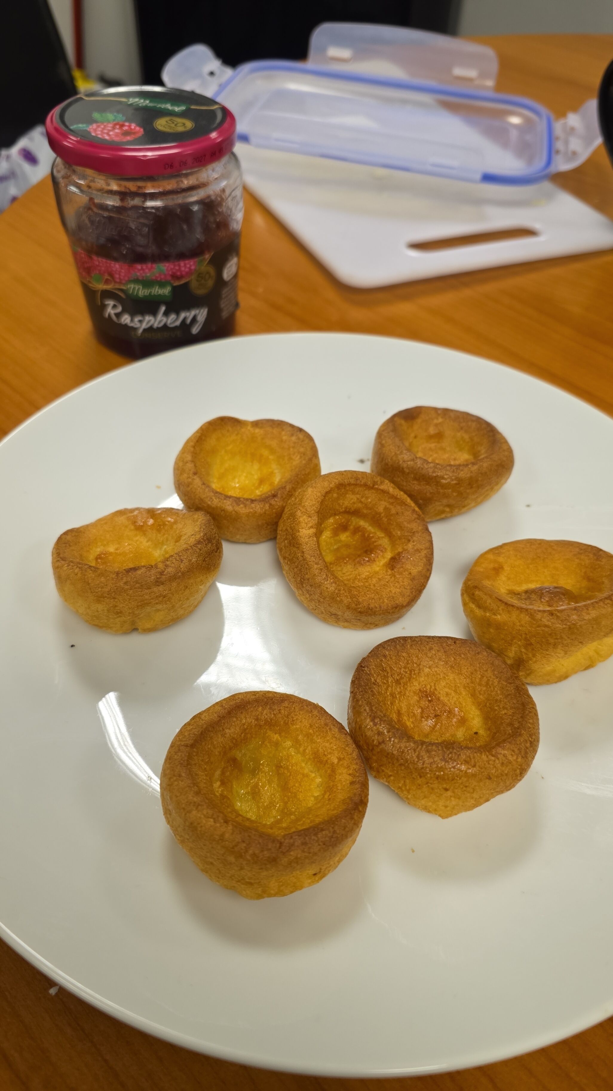 Une assiette de « Yorkshire puddings » sur une assiette blanche, avec un pot de confiture de framboises et un récipient en plastique en arrière-plan.