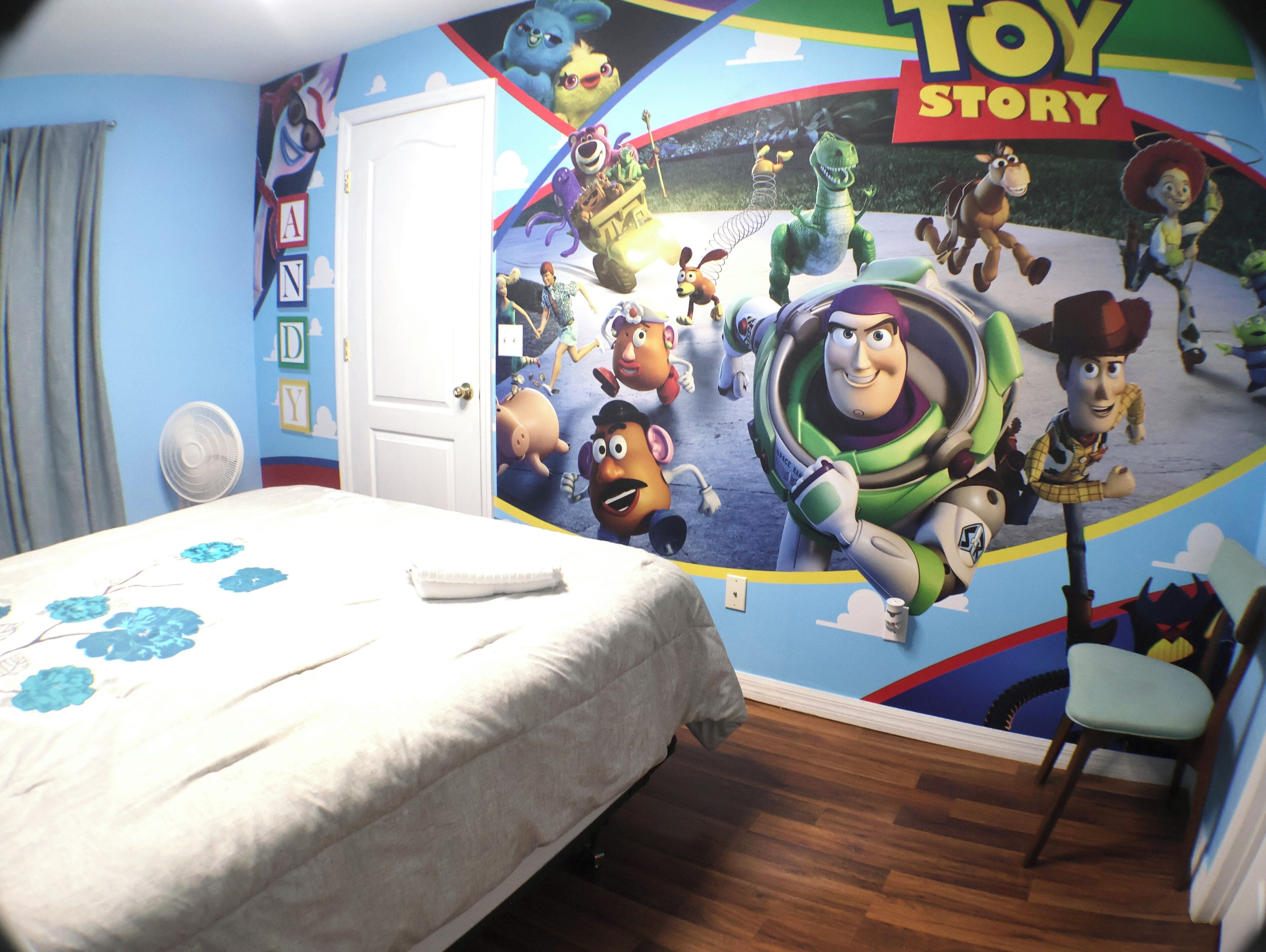 A bedroom with a bed, a chair, a wall mural of Toy Story characters, and a door.