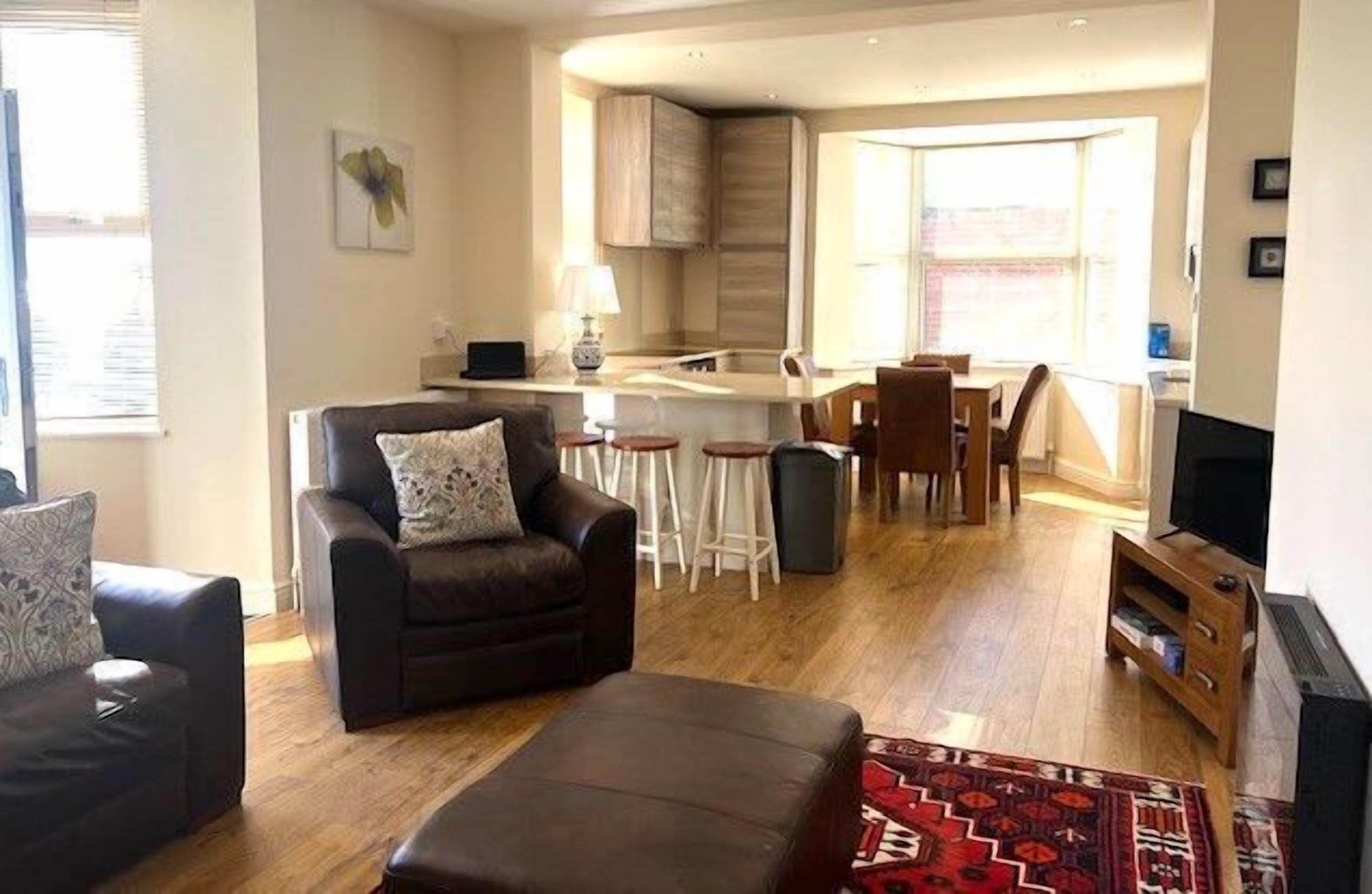A modern living room with a sofa, ottoman, and a TV stand. There is a kitchen area with a dining table and chairs.