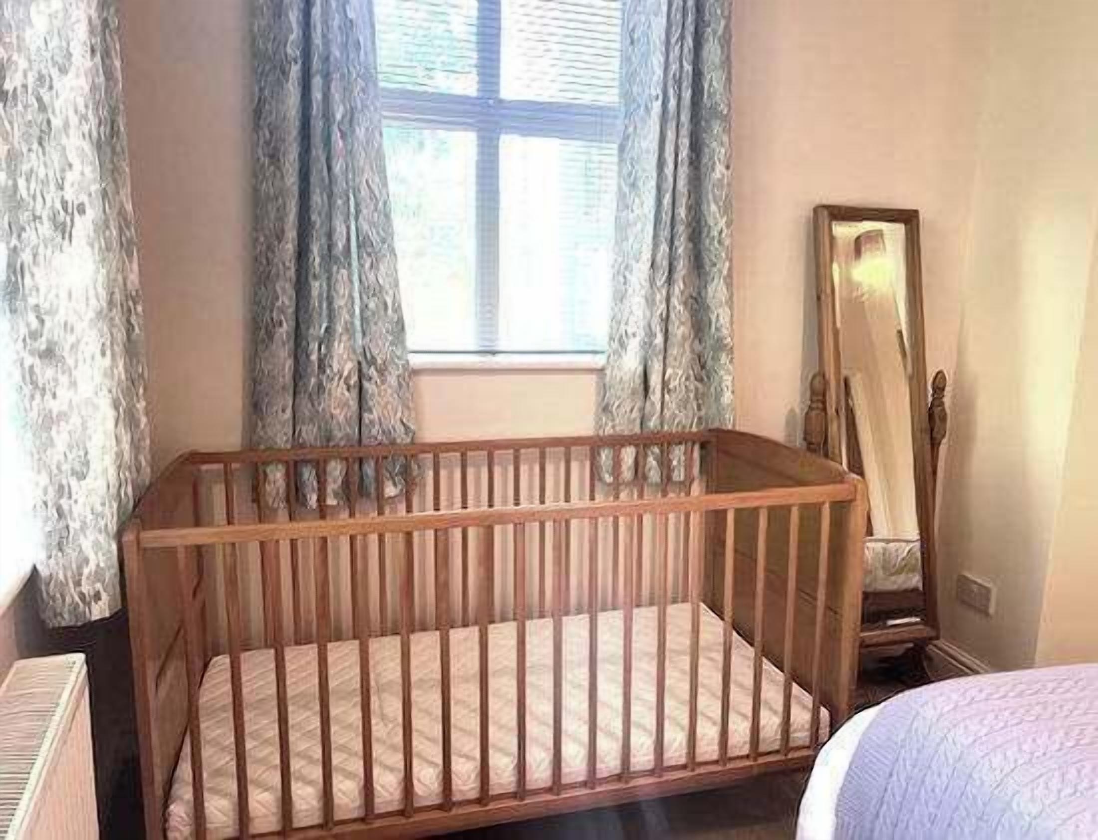 A wooden crib with a mattress, a window with curtains, a mirror, and a bed with a quilt.