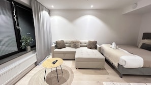 A modern bedroom with a sofa, a smaller bed, a round table, and a window with blinds.