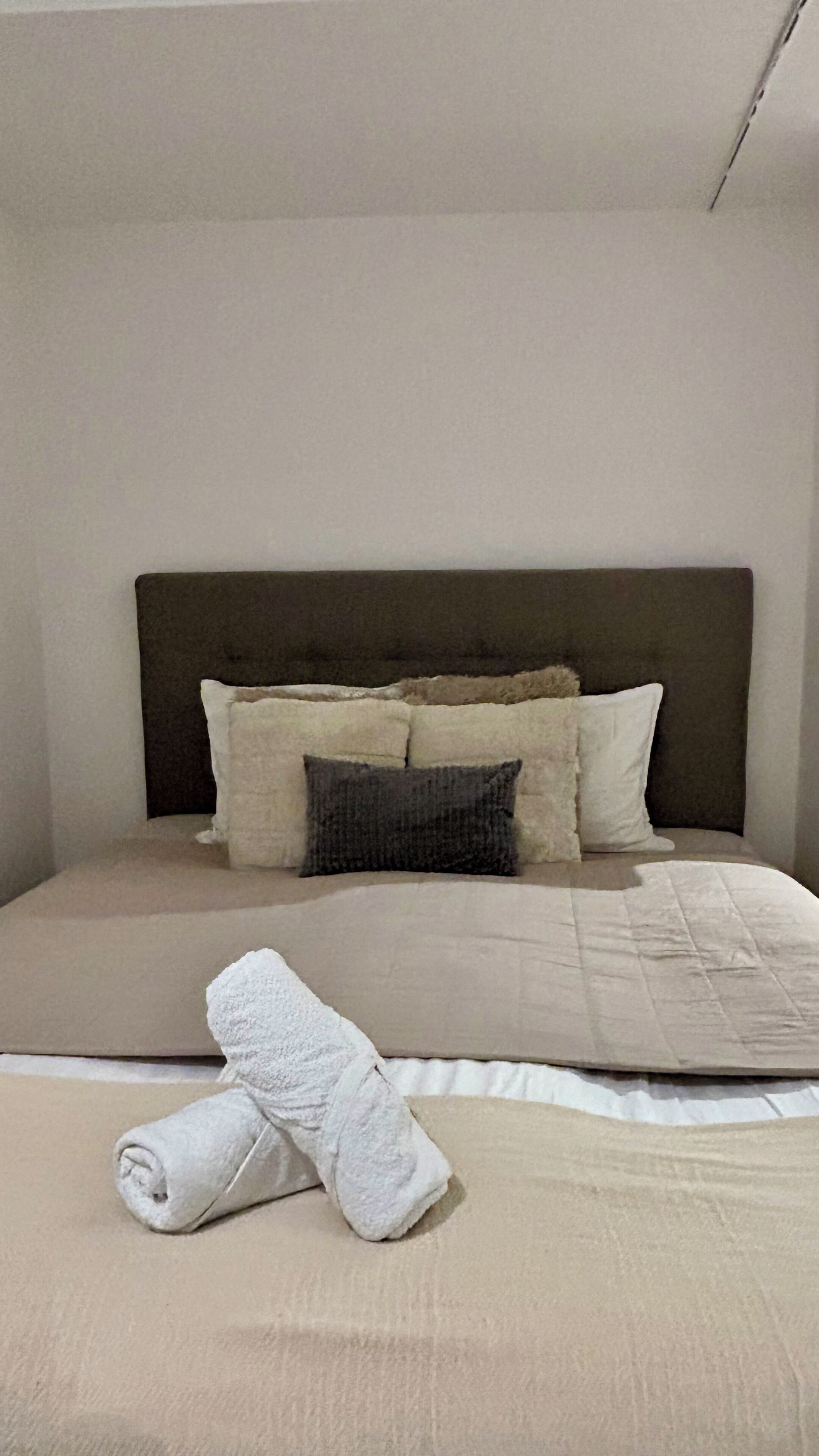 Un lit bien fait, avec une tête de lit sombre, plusieurs oreillers et une serviette blanche roulée posée sur le lit.