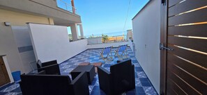 A rooftop patio with outdoor furniture, a view of the sea, and a modern building in the background. - The Turquoise Little House (Palinuro)