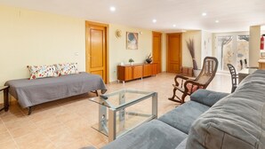 A living room with a sofa, a glass coffee table, a bed with patterned pillows, wooden cabinets, and a door leading to another room.