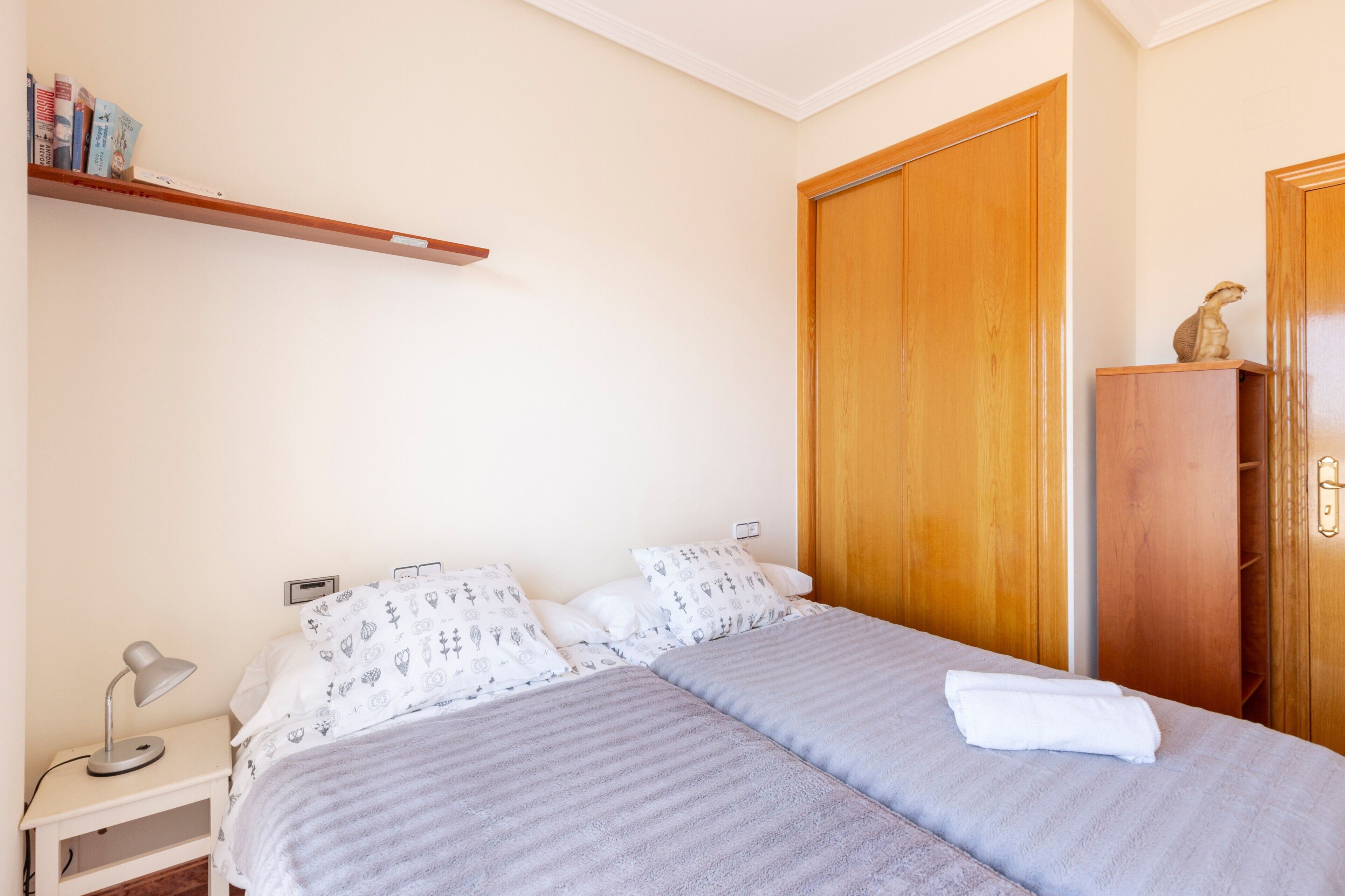 Een slaapkamer met twee bedden, een nachtkastje, een bureau, een houten kledingkast en een plank met boeken.