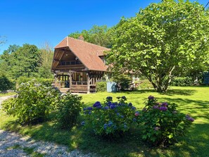 Een rustieke houten chalet met een schuin dak, omgeven door weelderige begroeiing en bloeiende planten.