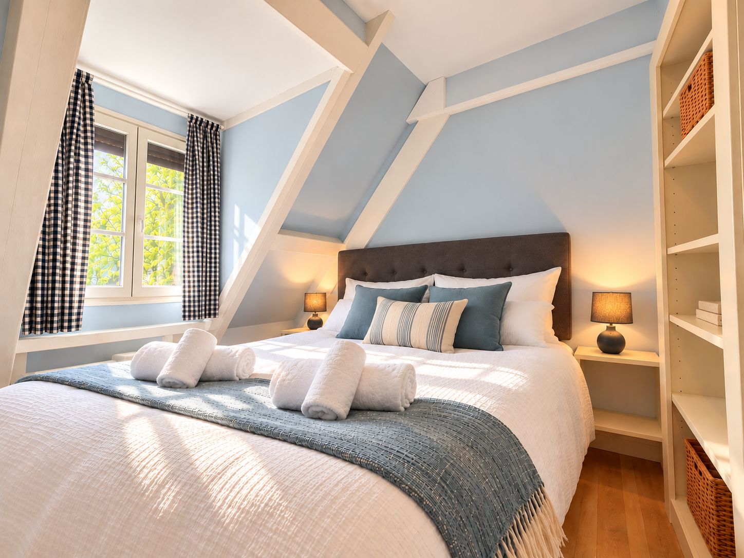 Ein Schlafzimmer mit schrägen Wänden, einem Bett mit weißer Bettwäsche, zwei Nachttischlampen und einem Fenster mit Vorhängen.