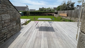 A wooden deck with a table and chairs, a stone wall, and a grassy yard with a hedge. - 3 bedroom house and garden facing the beach. Arzon. Gulf of Morbihan (Arzon)