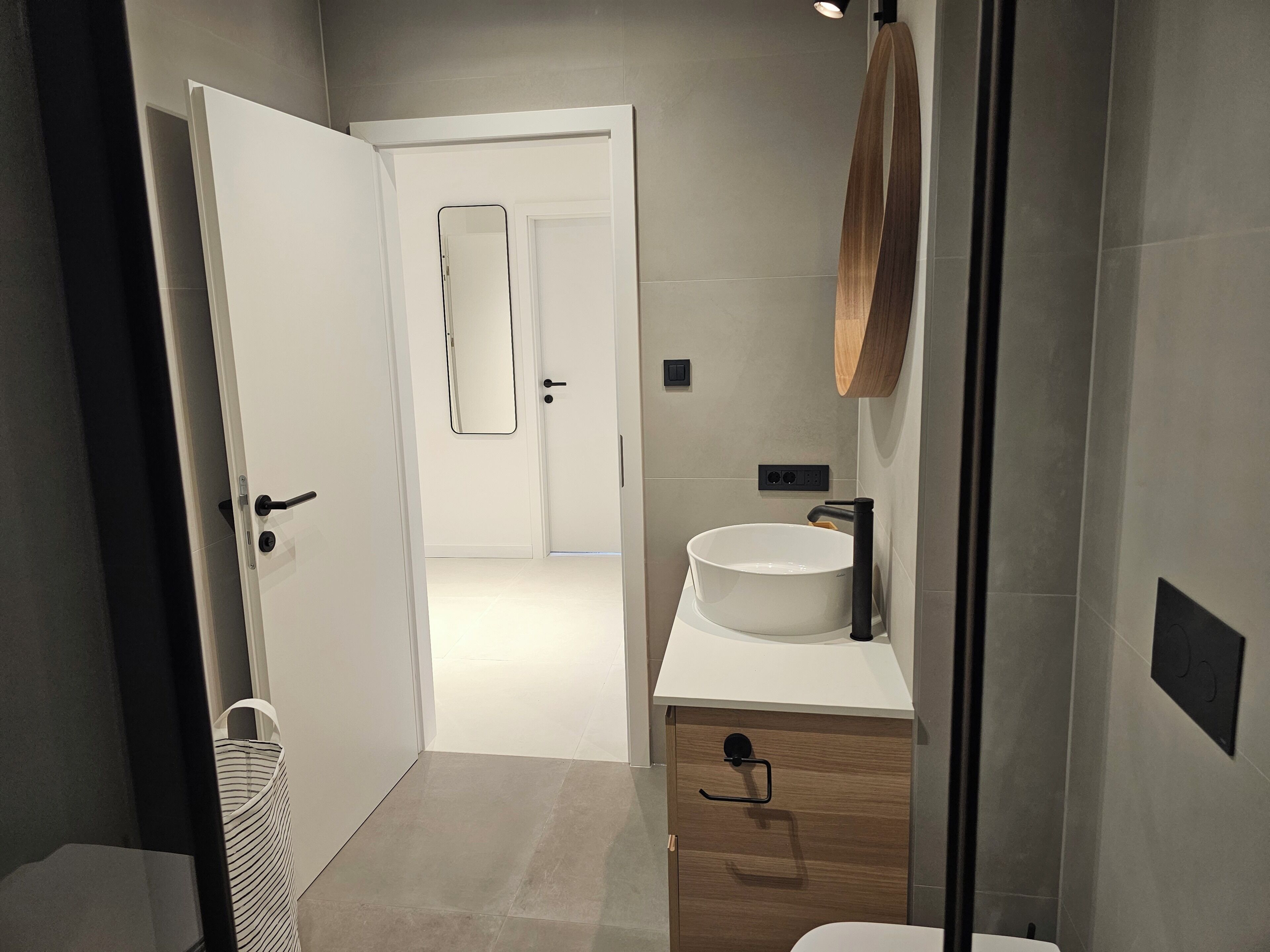 A modern bathroom with a white sink, wooden vanity, and a door leading to another room.