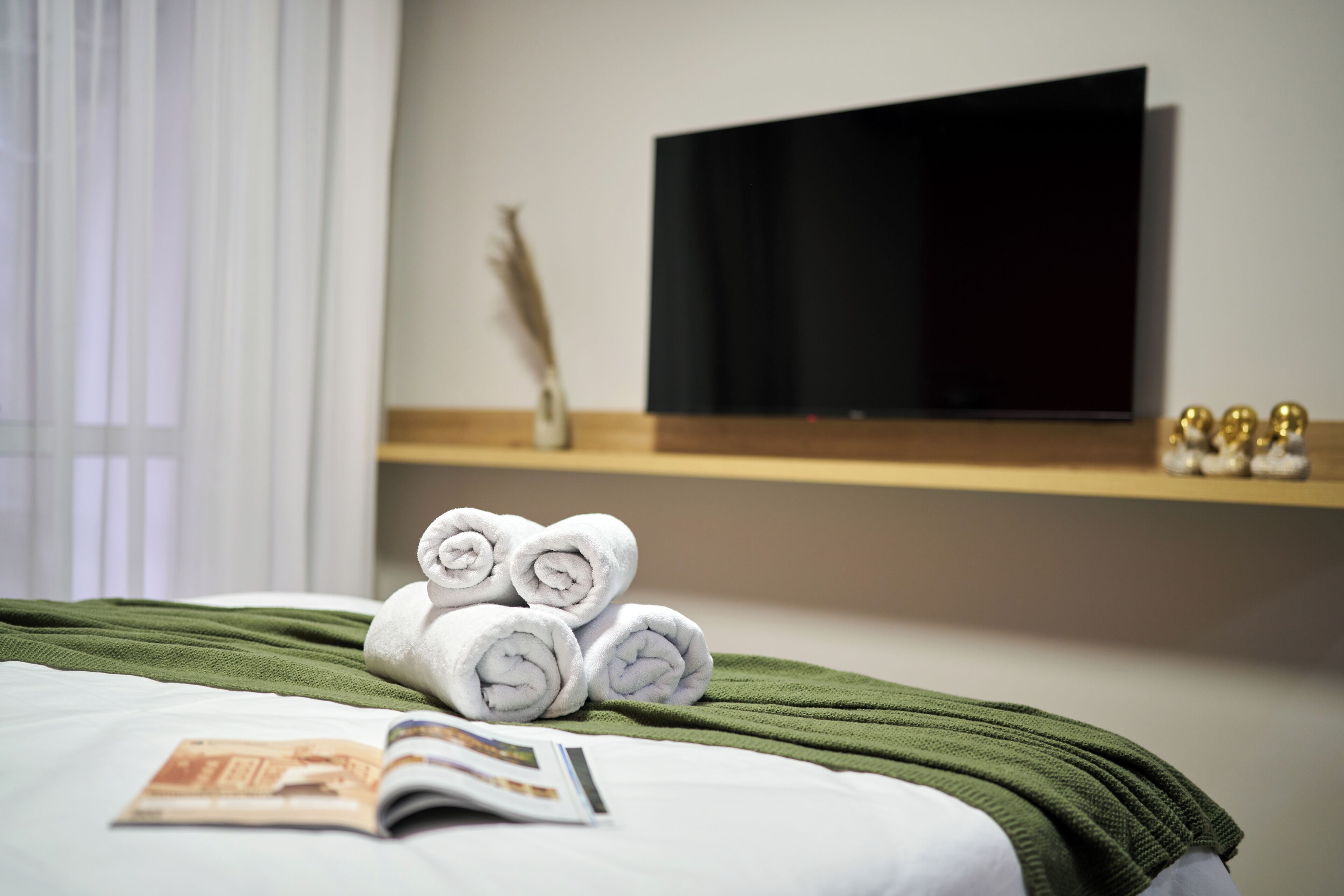 A neatly made bed with rolled towels, a green blanket, and a magazine on the bed.