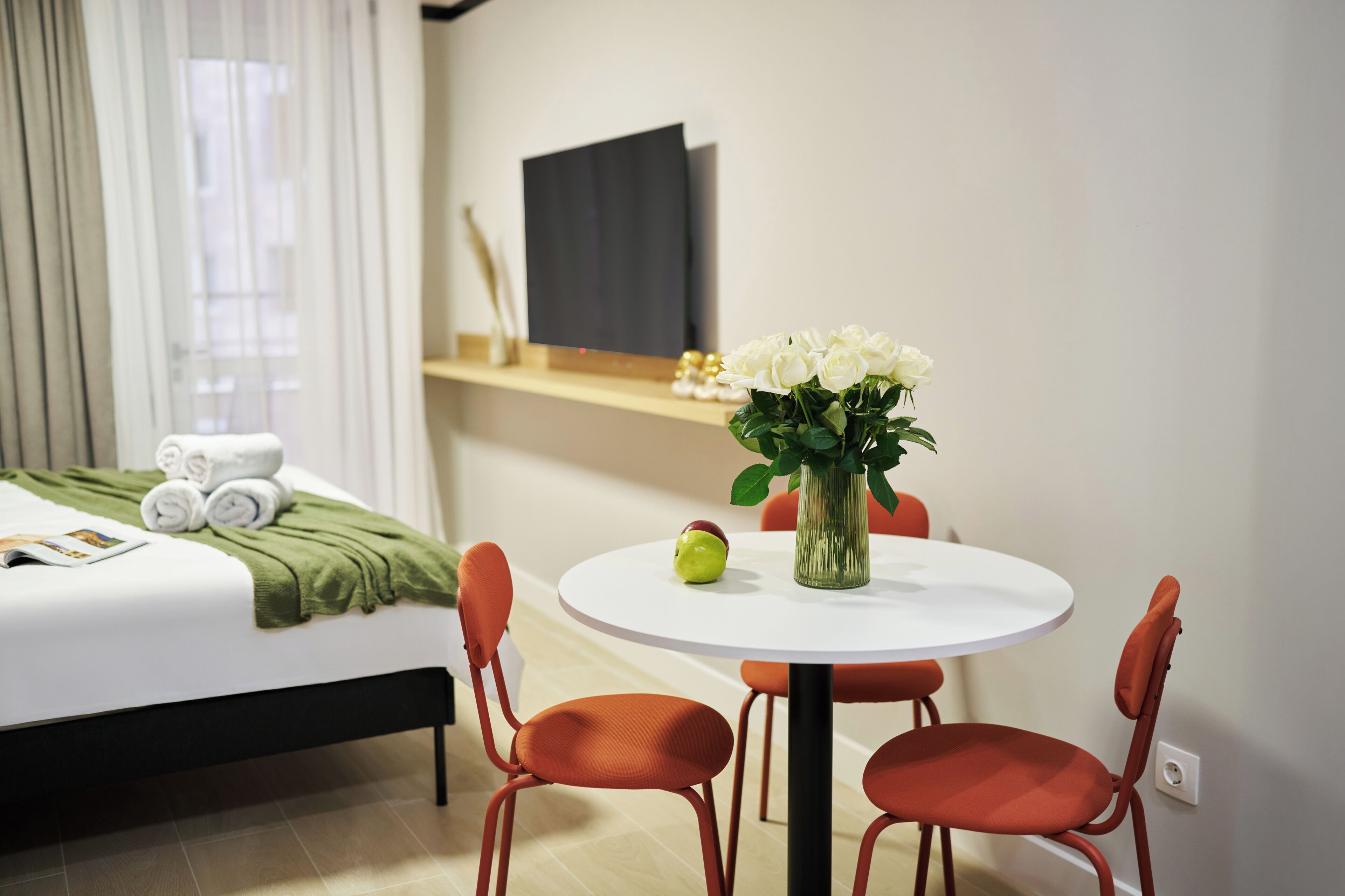 A hotel room with a bed, a round table with chairs, a vase with flowers, and a flat-screen TV on the wall.