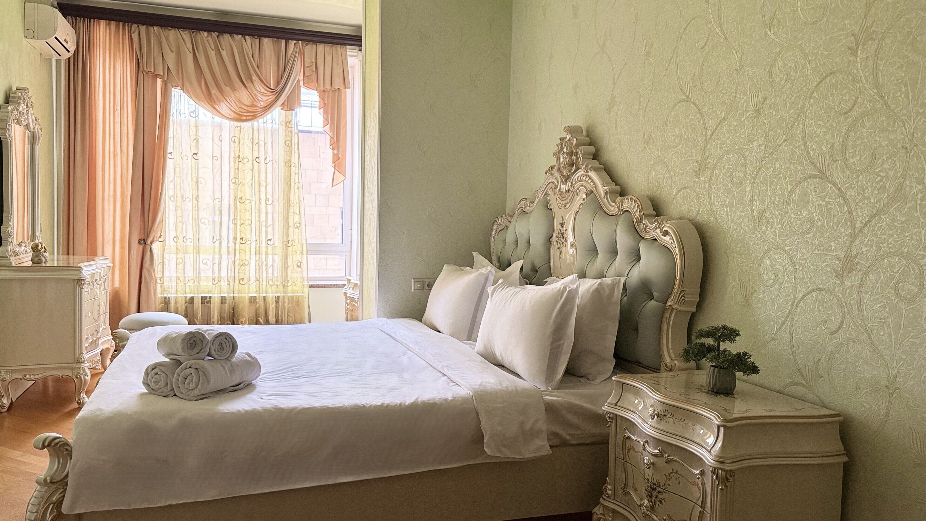 A neatly made bed with rolled towels, a classic headboard, and a small bedside table with a vase.