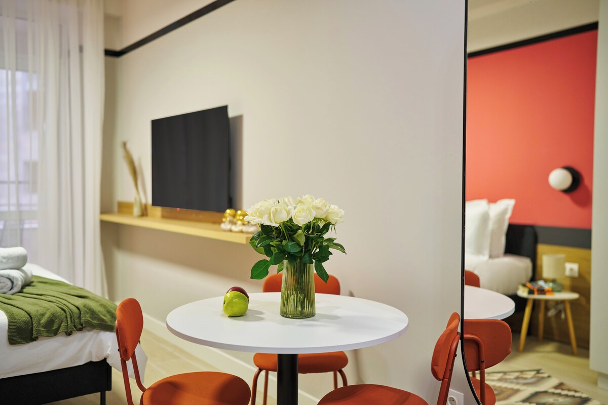 Une chambre d’hôtel moderne avec un lit, une table à manger ronde entourée de chaises, un vase de fleurs et un téléviseur à écran plat fixé au mur.
