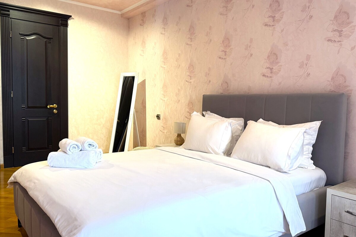 A neatly made bed with white linens, a grey headboard, and a bedside table with a lamp.