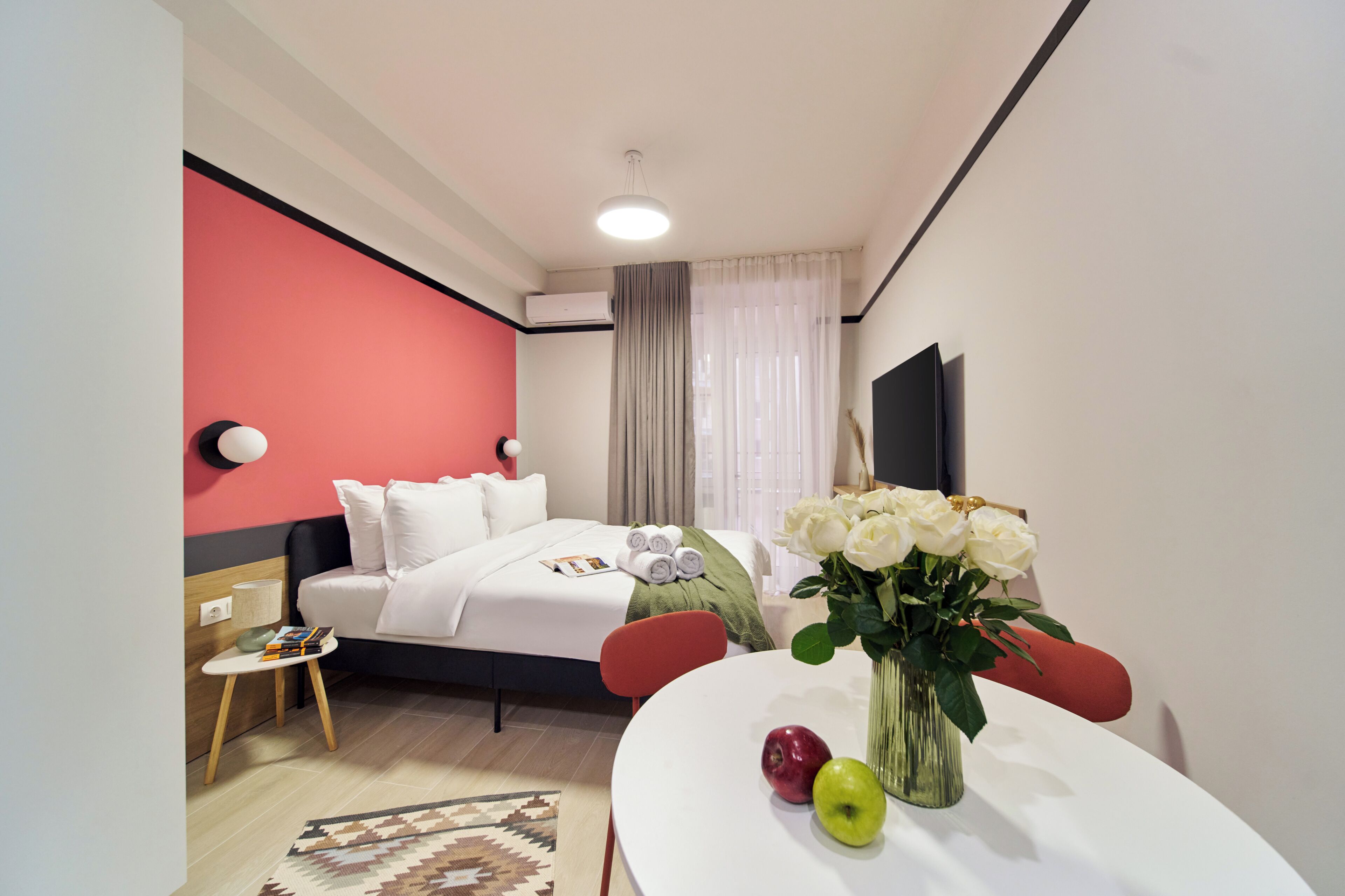 Una habitación de hotel con una cama, una mesa con manzanas y flores, un televisor y una pared roja.