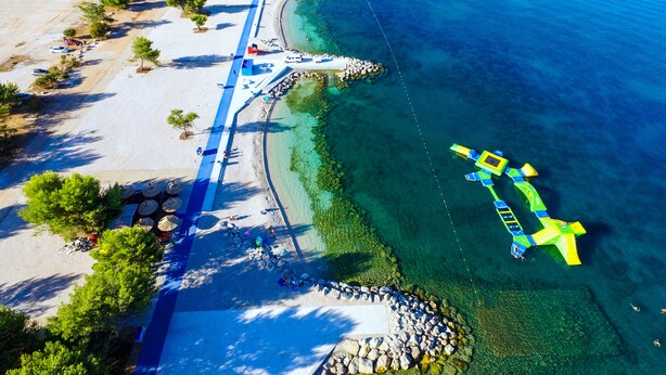 Vista aérea de una playa con mar azul cristalino, orilla arenosa y un paseo marítimo azul que se adentra en el agua.