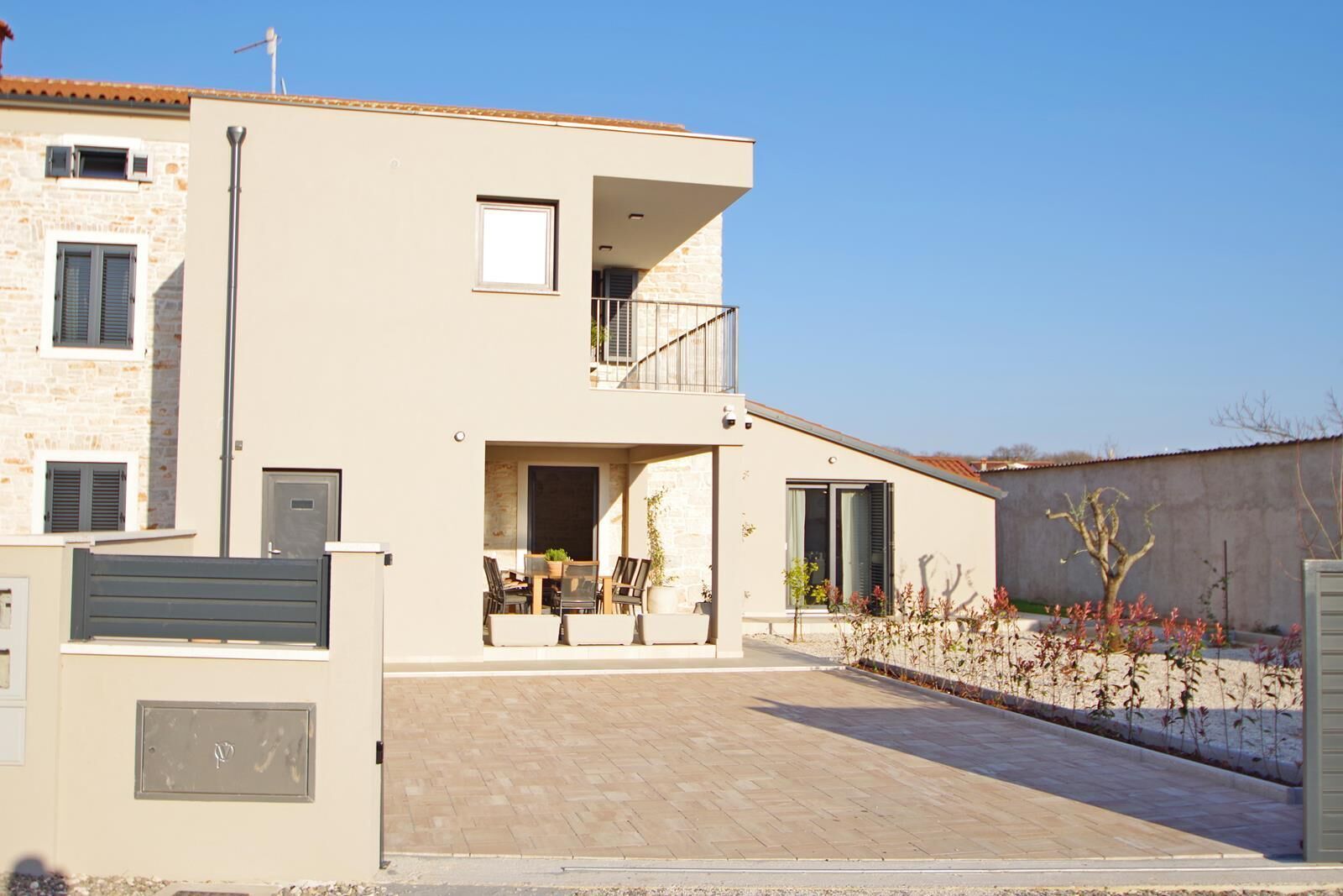 Une maison moderne à deux étages, dotée d’un balcon, d’une allée pavée et d’un jardin agrémenté d’arbustes.