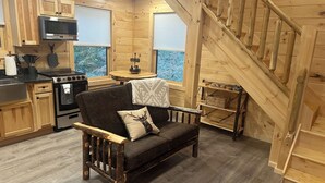 Interior acogedor de una cabaña con escalera de madera, sofá con un cojín de venado, área de cocina con estufa y microondas, y ventana con persianas.