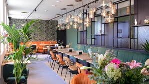 Een modern restaurant met binnenplanten, oranje stoelen en hangende verlichtingsarmaturen.