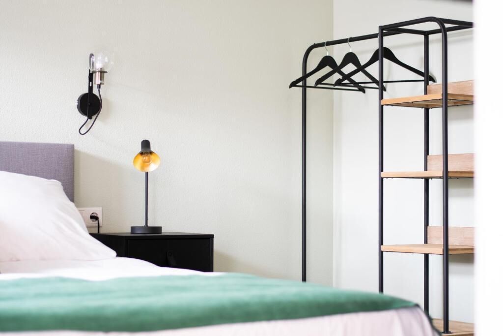 A bed with a green blanket, a bedside lamp, a wall-mounted light, and a black metal shelf unit.