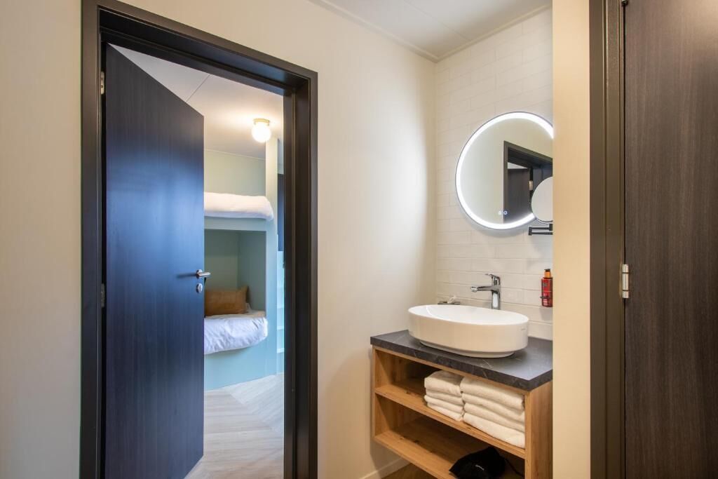 Un baño moderno con un espejo redondo, un lavamanos blanco y un estante de madera con toallas.