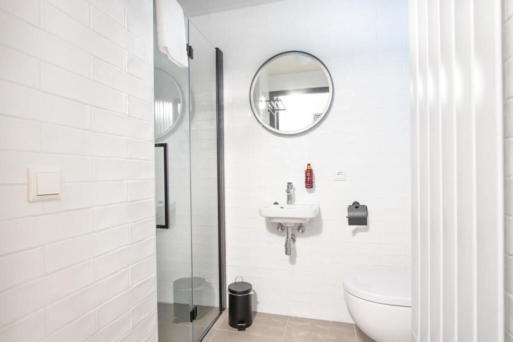 Un baño con paredes de azulejos blancos, un espejo redondo, un lavamanos, un inodoro y una cabina de ducha de vidrio.