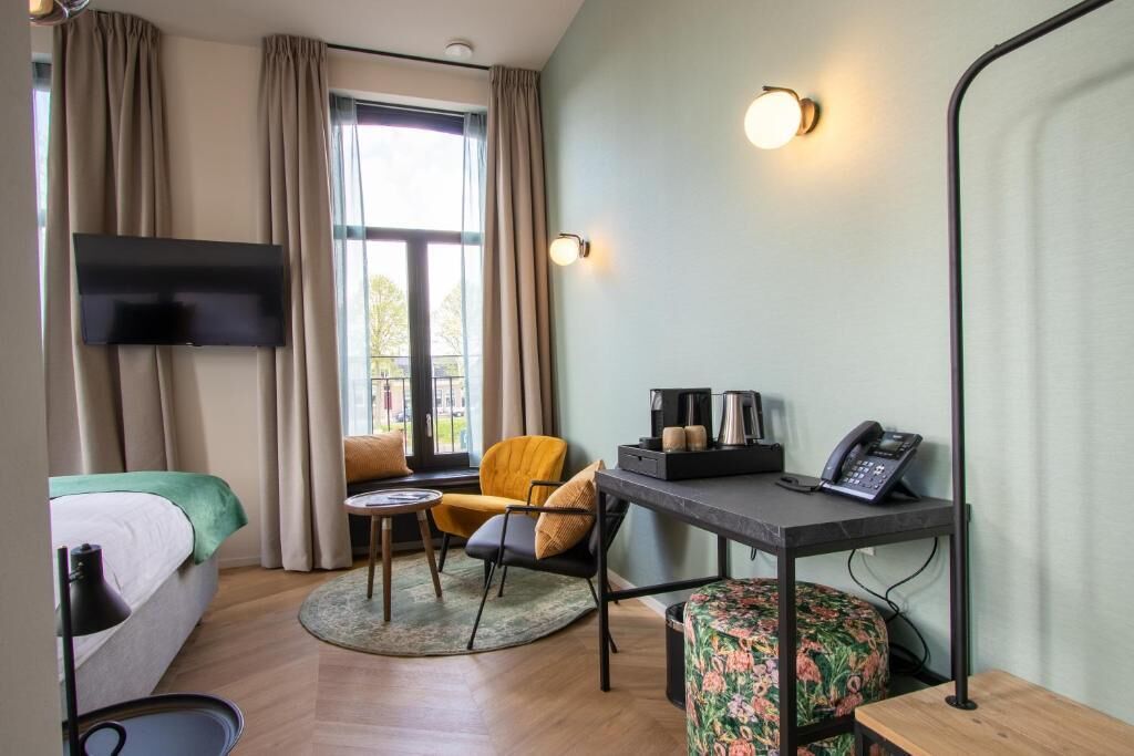 Ein Hotelzimmer mit einem Bett, einem Schreibtisch mit Telefon und Kaffeemaschine, einem Sessel, einem Fernseher und einem Fenster mit Vorhängen.