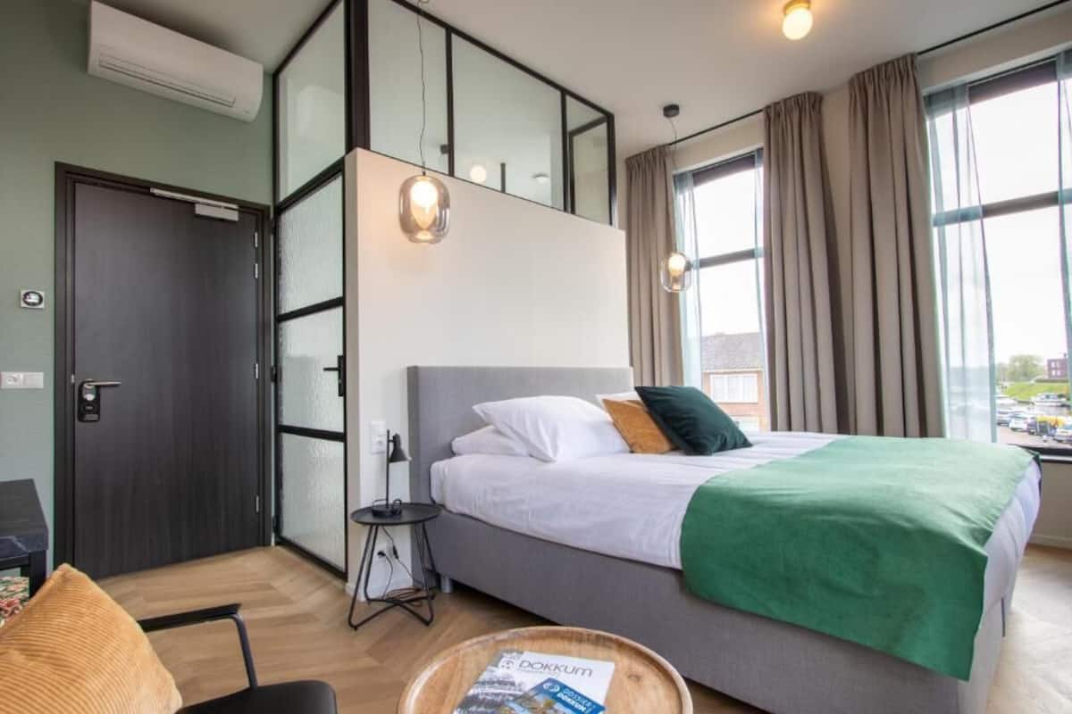 Ein Hotelzimmer mit einem Bett, einem kleinen Tisch mit einer Zeitschrift, einem Schreibtisch, einem Sessel, einem Fenster mit Vorhängen und einer Klimaanlage.