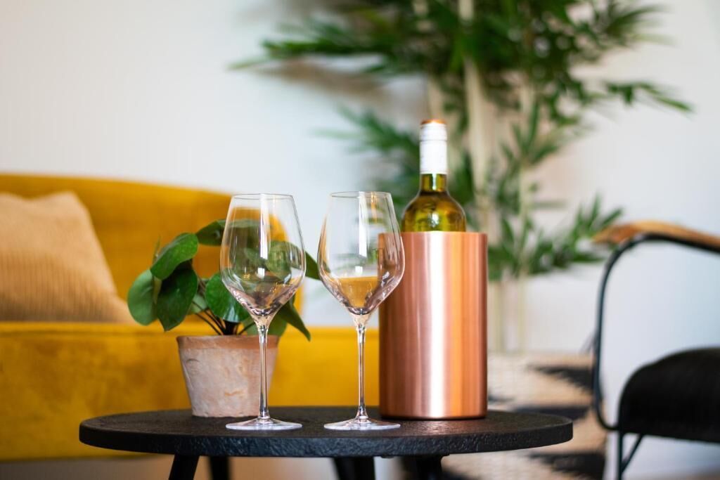 A table with two wine glasses and a copper decanter, a potted plant, and a yellow sofa in the background.